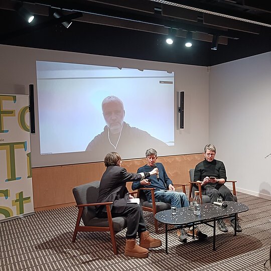 Three people seated on a stage with a large screen behind them showing a fourth person participating remotely.